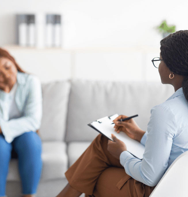 A therapist holds a clipboard and listens to a person sitting on a couch, who appears distressed and is leaning forward with their head resting on their hand. The setting is a bright, comfortable office.