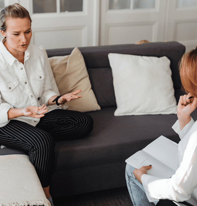 Two people sit on a gray couch having a serious conversation. One gestures with her hands while the other listens, holding a notebook and pen. The setting appears to be a casual living room or therapy session.