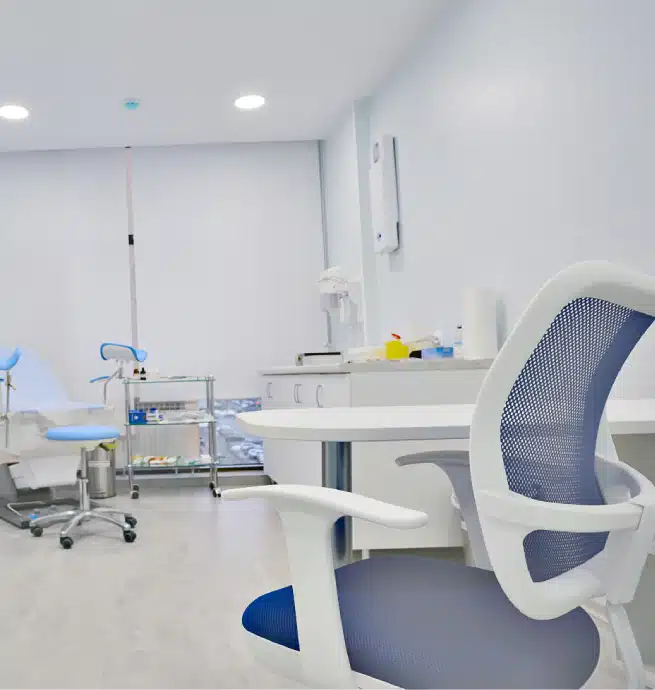 Modern medical examination room with a blue and white chair in the foreground, an examination table, medical equipment, and supplies on counters in a clean, brightly lit space.