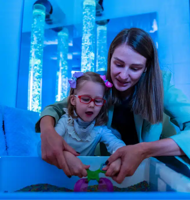A woman helps a young girl with glasses and bows in her hair play with a toy in a sensory room with blue lighting and bubble tubes in the background.