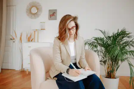 A woman with shoulder-length hair and glasses sits in a light-colored chair, writing in a notebook. She wears a beige blazer and dark pants. A large potted plant and modern decor are visible in the bright room.