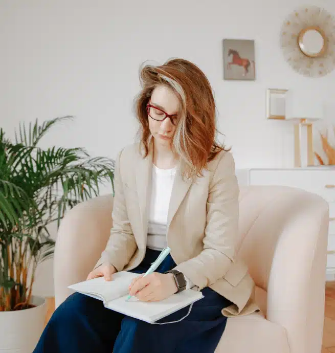 A woman with shoulder-length hair and glasses sits in a light pink armchair, writing in a notebook. She wears a beige blazer and dark pants. A plant and framed art are visible in the bright, modern room.
