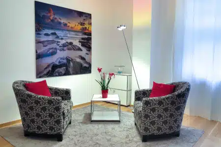 A cozy sitting area with two patterned armchairs, red cushions, a small table with a potted plant, a floor lamp, and a large wall art of a rocky beach scene, set on a light rug near sheer curtains.